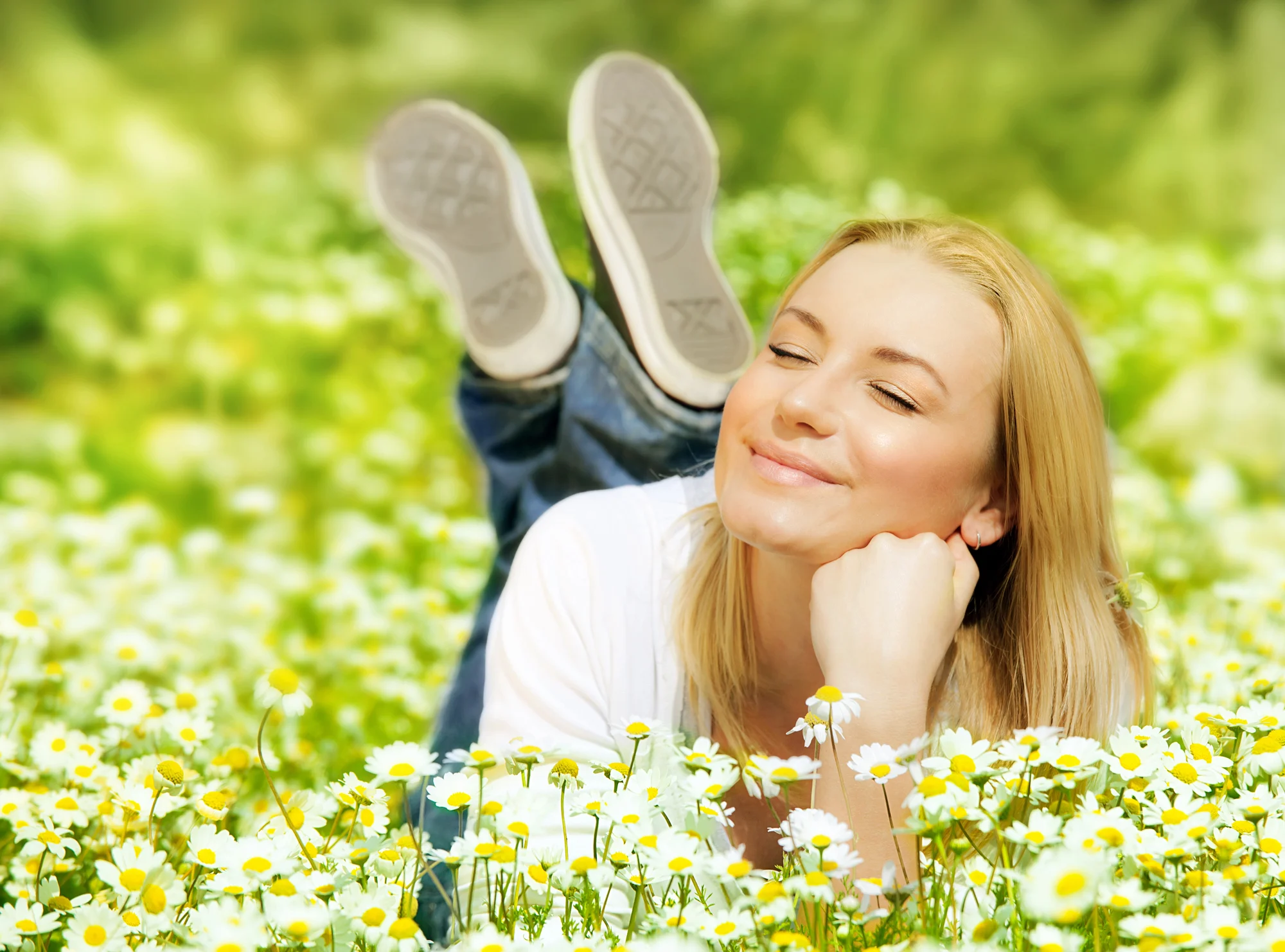 A woman with blonde hair lies in a field of daisies in Hazeldale, propped on her elbows with her feet raised behind her, smiling peacefully with her eyes closed under soft, natural sunlight.