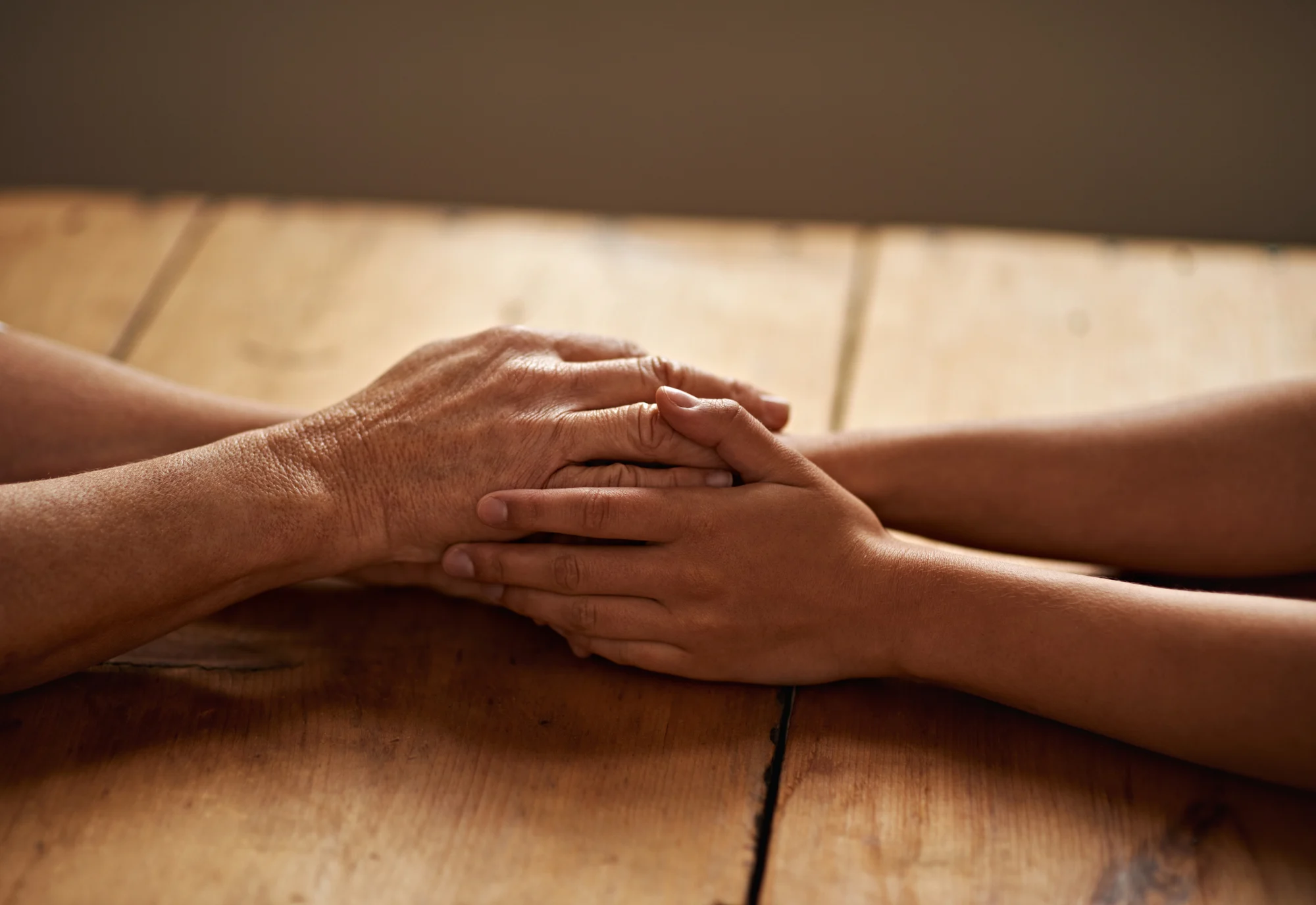 Two people sit at a wooden table, gently holding hands, conveying comfort, support, and connection—an atmosphere reminiscent of aloha. The photo focuses on their hands against a soft, neutral background.